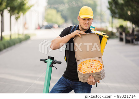 Happy smiling delivery man in yellow cap and thermal insulated bag offering pizza in cardboard box Happy smiling delivery man in yellow cap and thermal insulated bag offering pizza in cardboard box 89321714