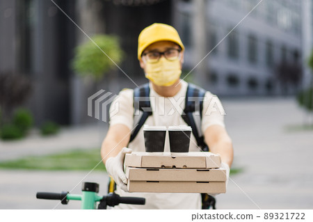 Delivery man in protective face mask holding stack of pizza cardboard boxes with two cups of coffee Delivery man in protective face mask holding stack of pizza cardboard boxes with two cups of coffee 89321722