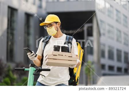 Courier in safety face mask, gloves and uniform holding pizza boxes and takeaway coffee cups 89321724