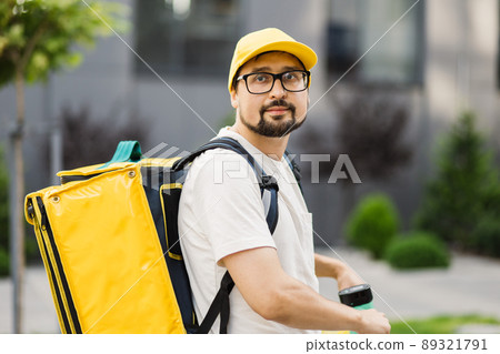 Young happy delivery man riding electric scooter with yellow thermal box backpack 89321791