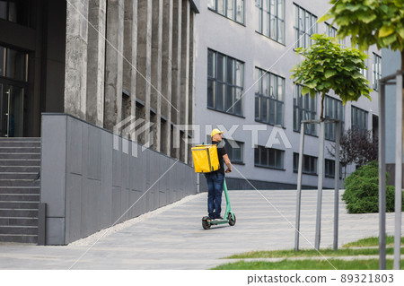 Young man with yellow backpack on his back in casual summer clothes is movingon an electric scooter Young man with yellow backpack on his back in casual summer clothes is movingon an electric scooter 89321803
