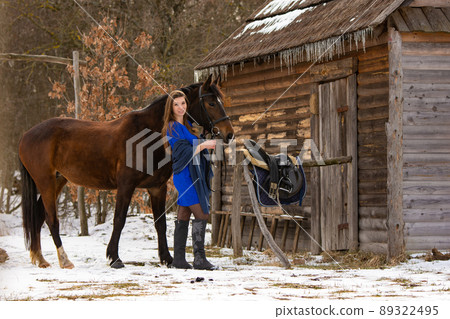 A beautiful girl in a short blue dress stands with a horse from an old wooden house A beautiful girl in a short blue dress stands with a horse from an old wooden house 89322495