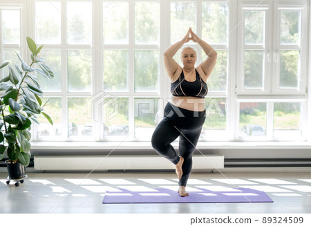 Plump chubby woman doing yoga standing in a tree pose. Female keeps balance by doing fitness while standing on a sports mat. Room with large high windows to the floor, letting in a lot of sunlight Plump chubby woman doing yoga standing in a tree pose. Female keeps balance by doing fitness while standing on a sports mat. Room with large high windows to the floor, letting in a lot of sunlight 89324509