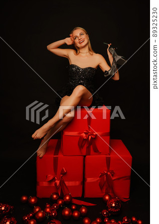 Beautiful blonde posing on a black background, wearing a black dress. Sits on red gift boxes. Red Christmas balls are scattered on the floor below. Christmas and New Year concept. 89325230
