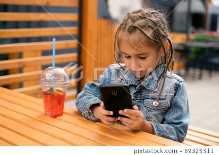 A child is sitting at a table on the outdoor terrace of a cafe and playing games on the phone. Participation in social networks. A girl in a denim jacket with pigtails looks at her smartphone A child is sitting at a table on the outdoor terrace of a cafe and playing games on the phone. Participation in social networks. A girl in a denim jacket with pigtails looks at her smartphone 89325311