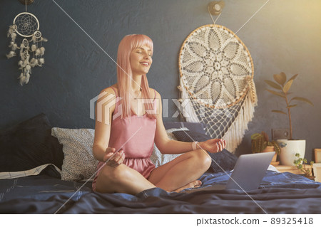 Pretty smiling female model meditates near laptop on large bed by dark blue wall Pretty smiling female model meditates near laptop on large bed by dark blue wall 89325418