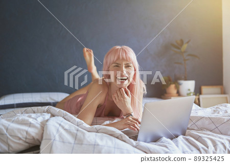 Attractive woman with bright pink laughs using modern laptop on bed by blue wall 89325425