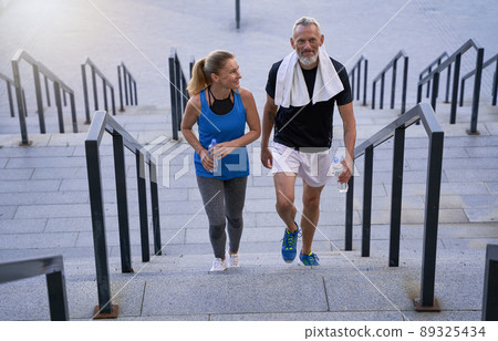Sporty mature couple, man and woman in sportswear looking pleased, walking up the stairs after training together outdoors Sporty mature couple, man and woman in sportswear looking pleased, walking up the stairs after training together outdoors 89325434