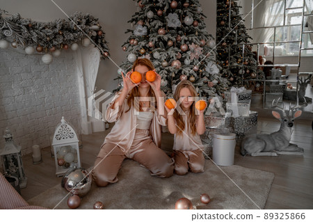 Mom and daughter hold tangerines at eye level instead of glasses. They are sitting under a decorated Christmas tree in their pajamas. The concept of Christmas, family holidays Mom and daughter hold tangerines at eye level instead of glasses. They are sitting under a decorated Christmas tree in their pajamas. The concept of Christmas, family holidays 89325866