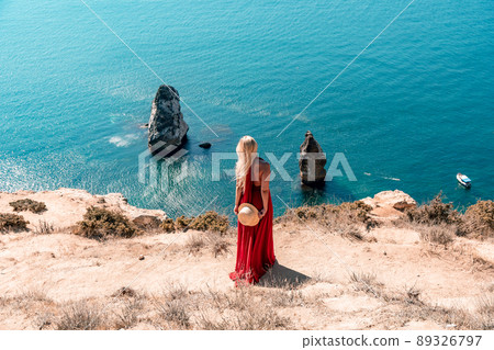 A girl with loose hair in a long red dress descends the stairs between the yellow rocks overlooking the sea. A rock can be seen in the sea. Sunny path on the sea from the rising sun A girl with loose hair in a long red dress descends the stairs between the yellow rocks overlooking the sea. A rock can be seen in the sea. Sunny path on the sea from the rising sun 89326797