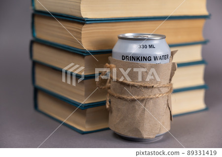 A stack of books and a can of drinking water against a gray background. Blue hardcover books. Can of freshwater wrapped in paper and tied with string. A stack of books and a can of drinking water against a gray background. Blue hardcover books. Can of freshwater wrapped in paper and tied with string. 89331419