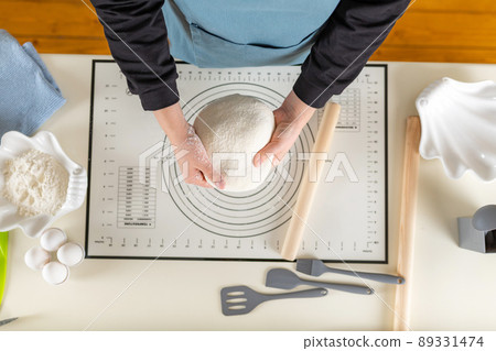 The hands of a cook in a blue apron hold wheat dough over a kitchen table, top view, selective focus The hands of a cook in a blue apron hold wheat dough over a kitchen table, top view, selective focus 89331474