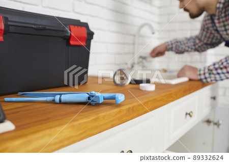 Plumber using a wrench and other tools while repairing the sink tap in the kitchen Plumber using a wrench and other tools while repairing the sink tap in the kitchen 89332624