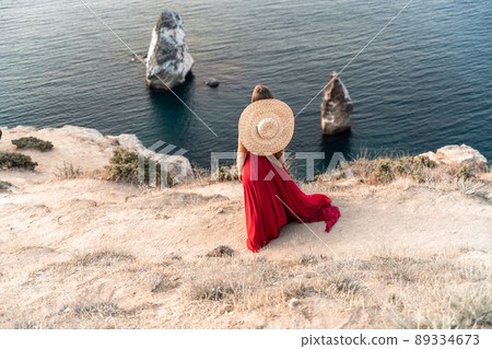 A girl with loose hair in a red dress and hat stands on the rocks above the sea. In the background, the sea and the rocks. The concept of travel. 89334673