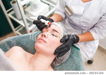Doctor cosmetologist makes facial massage procedure using a dermo roller. Woman in beauty salon during mesotherapy procedure with mesoscooter.. Doctor cosmetologist makes facial massage procedure using a dermo roller. Woman in beauty salon during mesotherapy procedure with mesoscooter.. 89335184