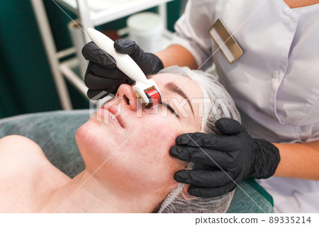 Doctor cosmetologist makes facial massage procedure using a dermo roller. Woman in beauty salon during mesotherapy procedure with mesoscooter Doctor cosmetologist makes facial massage procedure using a dermo roller. Woman in beauty salon during mesotherapy procedure with mesoscooter 89335214