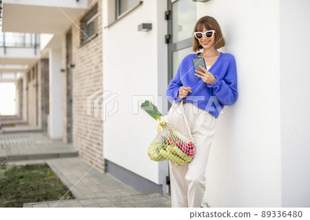 Woman with phone and mesh bag full of fresh vegetables on porch of home 89336480