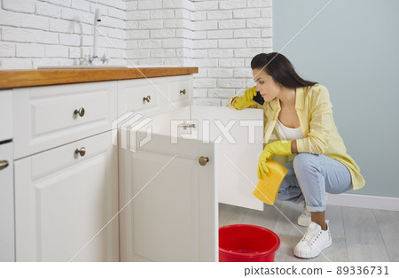 Frustrated woman on phone calls plumber because of leaking pipe in sink in kitchen. Frustrated woman on phone calls plumber because of leaking pipe in sink in kitchen. 89336731