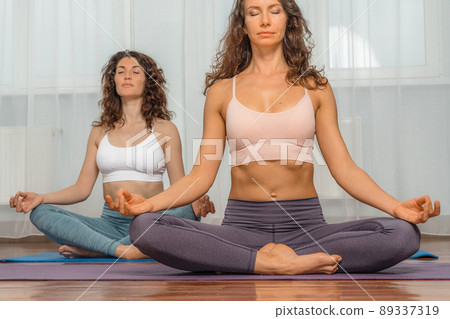 Two attractive athletic girls train asana yoga pose on mats on a light background. A group of young women stretch out in the gym. Healthy lifestyle concept Two attractive athletic girls train asana yoga pose on mats on a light background. A group of young women stretch out in the gym. Healthy lifestyle concept 89337319