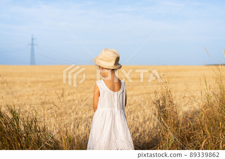 Little girl standing yellow wheat field dressed white dress and straw hat Little girl standing yellow wheat field dressed white dress and straw hat 89339862