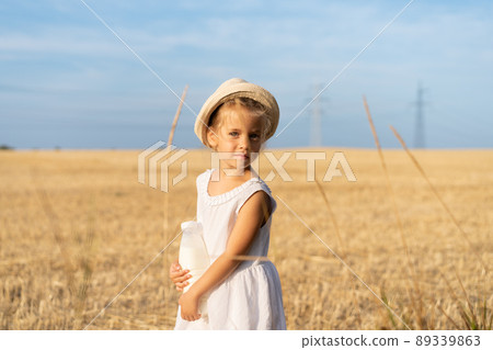 Little girl standing wheat field holding bottle of milk in hand dressed white dress and straw hat Little girl standing wheat field holding bottle of milk in hand dressed white dress and straw hat 89339863