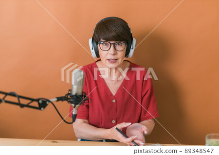 Mature woman making podcast recording for her online show. Attractive business woman using headphones front of microphone for a radio broadcast 89348547