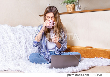 Home office woman and distance learning online education. Happy lady girl with wavy hair working at notebook sit down on bed and drinking coffee at home. Using computer and online shops. Stay at home Home office woman and distance learning online education. Happy lady girl with wavy hair working at notebook sit down on bed and drinking coffee at home. Using computer and online shops. Stay at home 89350198