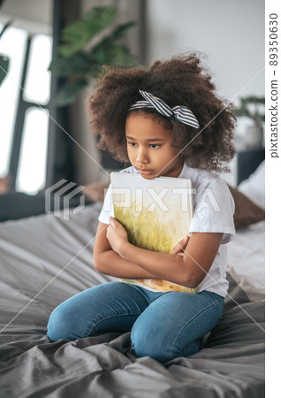 A girl sigtting on a ned with a book in hands and looking thoughful 89350630