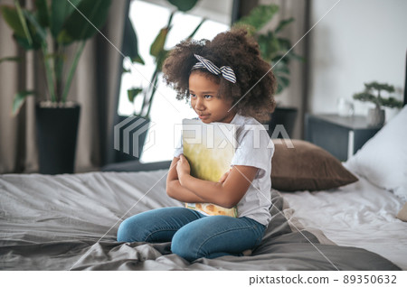 A girl sigtting on a ned with a book in hands and looking thoughful A girl sigtting on a ned with a book in hands and looking thoughful 89350632