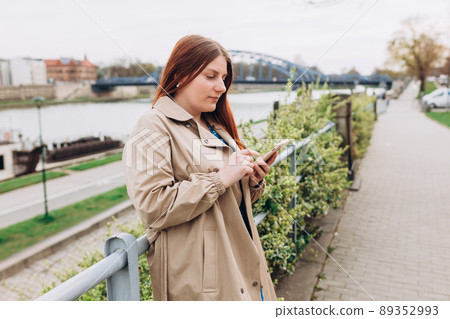 Beautiful happy redhead woman is using a smart phone and smiling outdoors. Urban lifestyle concept. Girl walking on city street checks her smartphone. Spring time 89352993