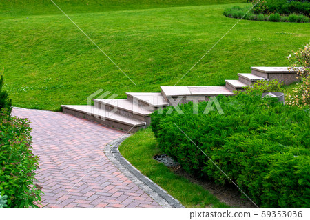 Granite steps to path from brick stone tile in a park on a slope with evergreen bushes and turf lawn, on garden bed lattice drain system and iron manhole, nobody. Granite steps to path from brick stone tile in a park on a slope with evergreen bushes and turf lawn, on garden bed lattice drain system and iron manhole, nobody. 89353036