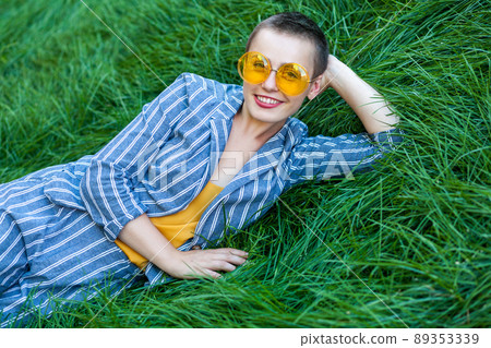 Portrait of beautiful young woman with short hair in casual blue striped suit, yellow shirt and glasses lying down on green grass, relax, looking at camera with toothy smile. outdoor summertime shot. 89353339