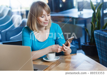 Top view portrait of young happy blonde woman in blue t-shirt, sitting in cafe and holding her mobile smart phone and texting. indoor studio shot. 89353353