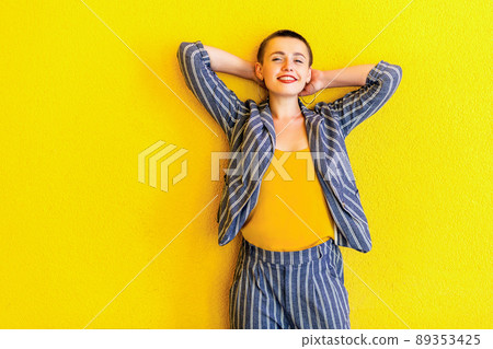Portrait of happy young short hair beautiful woman in yellow shirt and striped suit standing, holding her head and looking at camera with toothy smile. indoor studio shot isolated on yellow background 89353425