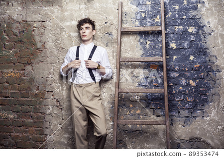 fashionable man in white shirt and beige pants holding suspenders posing and lean on old wooden ladder on brick wall background. indoor, studio shot. fashionable man in white shirt and beige pants holding suspenders posing and lean on old wooden ladder on brick wall background. indoor, studio shot. 89353474