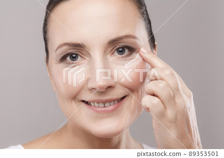 Middle aged woman with perfect face skin applying cosmetic cream on skin near eyes and looking at camera with toothy smile isolated on grey background. Skin care concept,copy space,studio shot, indoor 89353501