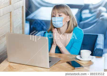 please help. Portrait of worry young woman with surgical medical mask is sitting and working on laptop and looking display and begging on video call. Indoor working, medicine and health care concept. 89353512