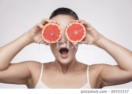 Closeup portrait of suprized woman with perfect skin smiling after cream, balm, mask, lotion, holding half of grapefruit and covering her eye, isolated on grey background. Indoor, studio shot. Closeup portrait of suprized woman with perfect skin smiling after cream, balm, mask, lotion, holding half of grapefruit and covering her eye, isolated on grey background. Indoor, studio shot. 89353537