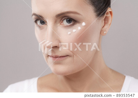 Middle aged woman with perfect face skin applying cosmetic cream on skin near eyes and looking at camera isolated on grey background. Beauty and skin care concept, copy space, studio shot, indoor Middle aged woman with perfect face skin applying cosmetic cream on skin near eyes and looking at camera isolated on grey background. Beauty and skin care concept, copy space, studio shot, indoor 89353547