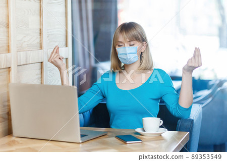 Portrait of calm young blonde woman with surgical medical mask in blue t-shirt is sitting and working on laptop and try to relax in yoga pose. Indoor working, medicine and health care concept. Portrait of calm young blonde woman with surgical medical mask in blue t-shirt is sitting and working on laptop and try to relax in yoga pose. Indoor working, medicine and health care concept. 89353549