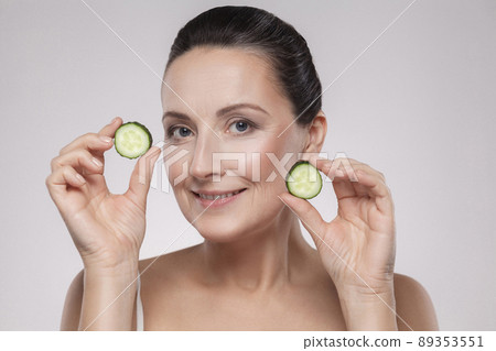 Natural spa treatment. Portrait of beautiful happy mature woman holding pieces of cucumber, showing and smiling while standing isolated on grey background. Copy space, indoor, studio shot 89353551