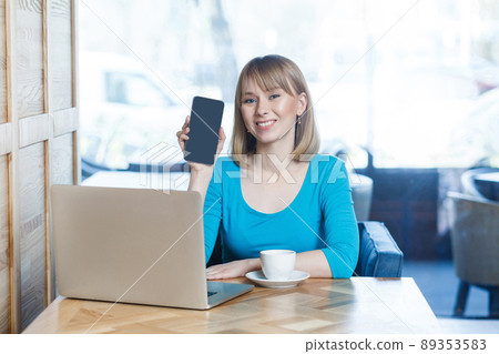 Portrait of happy beautiful young blonde woman in blue t-shirt, sitting with laptop, holding and showing mobile display and looking at camera with toothy smile. indoor, studio shot. Portrait of happy beautiful young blonde woman in blue t-shirt, sitting with laptop, holding and showing mobile display and looking at camera with toothy smile. indoor, studio shot. 89353583