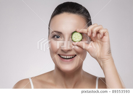 Sensitive care for luminous skin. Attractive middle aged woman looking at camera and covering eye with slice of cucumber while standing against grey background. Copy space, indoor,studio shot,isolated 89353593