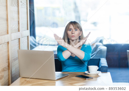 Enough! Portrait of warning aggressive young girl with blonde hair in t-shirt blouse are sitting in cafe and working on laptop and showing crossing raised arms like stop gesture. Indoor Enough! Portrait of warning aggressive young girl with blonde hair in t-shirt blouse are sitting in cafe and working on laptop and showing crossing raised arms like stop gesture. Indoor 89353602