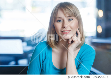 Portrait of beautiful happy attravctive blonde young woman in blue t-shirt with makeup and bangs hair sitting, touching her face and looking at camera with satisfied face. indoor, studio shot. Portrait of beautiful happy attravctive blonde young woman in blue t-shirt with makeup and bangs hair sitting, touching her face and looking at camera with satisfied face. indoor, studio shot. 89353605