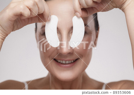 Patch Under Eyes. Beautiful middle aged woman with perfect skin showing white hydrogel patches with lifting anti-wrinkle collagen effect, gray background, indoor, studio shot, copy space, isolated 89353606