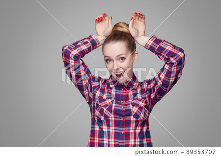 Funny beautiful blonde girl with pink checkered shirt, collected bun hairstyle and makeup standing and looking at camera with bunny gesture hands. indoor studio shot, isolated on gray background. Funny beautiful blonde girl with pink checkered shirt, collected bun hairstyle and makeup standing and looking at camera with bunny gesture hands. indoor studio shot, isolated on gray background. 89353707