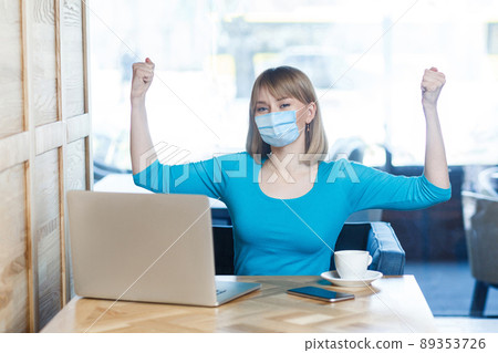 I am strong. Portrait of young woman with surgical medical mask in blue t-shirt is sitting and looking at camera and showing her biceps and satisfied. Indoor working, medicine and health care concept. I am strong. Portrait of young woman with surgical medical mask in blue t-shirt is sitting and looking at camera and showing her biceps and satisfied. Indoor working, medicine and health care concept. 89353726
