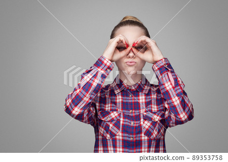 beautiful funny blonde girl in red, pink checkered shirt, collected bun hairstyle, makeup standing and looking at camera with binoculars gesture hands. indoor studio shot. isolated on gray background 89353758