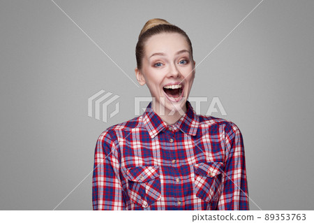 funny beautiful blonde girl in red, pink checkered shirt, collected bun hairstyle, makeup standing and looking at camera and laughing with open mouth. indoor studio shot. isolated on gray background funny beautiful blonde girl in red, pink checkered shirt, collected bun hairstyle, makeup standing and looking at camera and laughing with open mouth. indoor studio shot. isolated on gray background 89353763